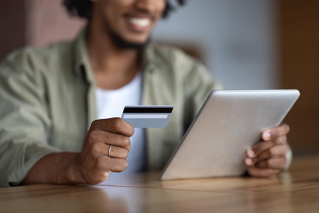 Ragazzo afroamericano felice millenario ritagliato con tablet seduto al tavolo e tenere la carta di credito