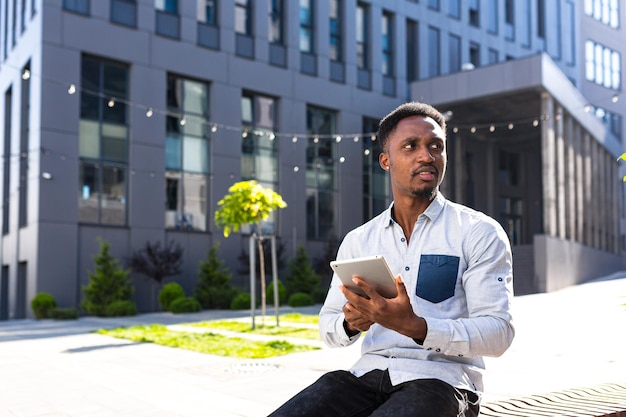 Ragazzo afroamericano utilizzando tablet seduto sulla panchina sulla strada della città su sfondo urbano. L'uomo di colore legge o fa acquisti tenendo in mano un gadget o uno smartphone. uno studente del campus. libero professionista fuori