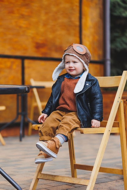 Ragazzo alla moda di 3 anni con una giacca di pelle e pantaloni marroni si siede sulla terrazza vicino al caffè Bambino moderno Moda per bambini Bambino felice