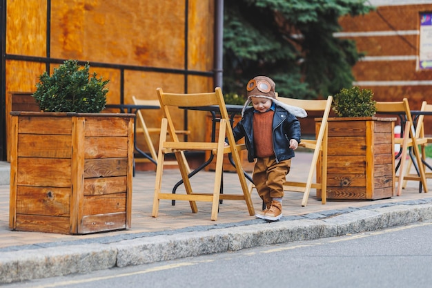 Ragazzo alla moda di 3 anni con una giacca di pelle e pantaloni marroni si siede sulla terrazza vicino al caffè Bambino moderno Moda per bambini Bambino felice