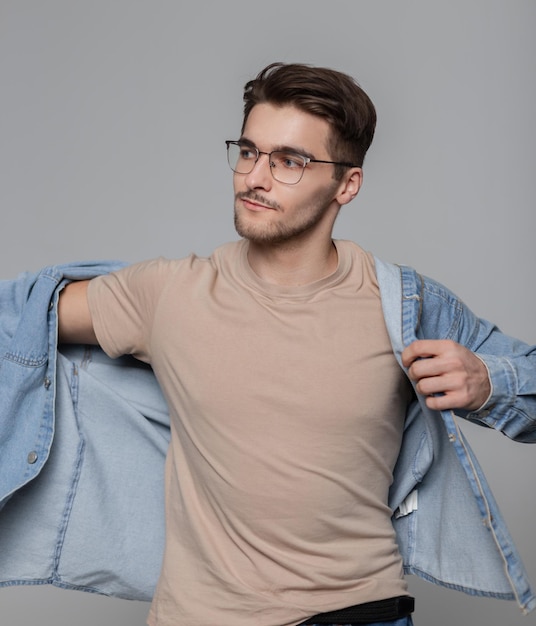 Ragazzo alla moda modello hipster americano con acconciatura e occhiali vintage in abiti alla moda con una maglietta beige che indossa una camicia di jeans su sfondo grigio