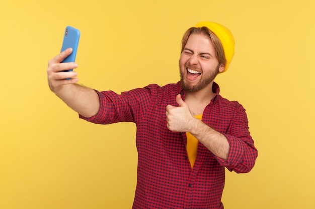 Ragazzo allegro alla moda hipster in camicia a scacchi che mostra pollice in alto gesto di approvazione sorridente parlando videochiamata o streaming sul cellulare chiedendo di seguire e come il suo blog girato in studio isolato