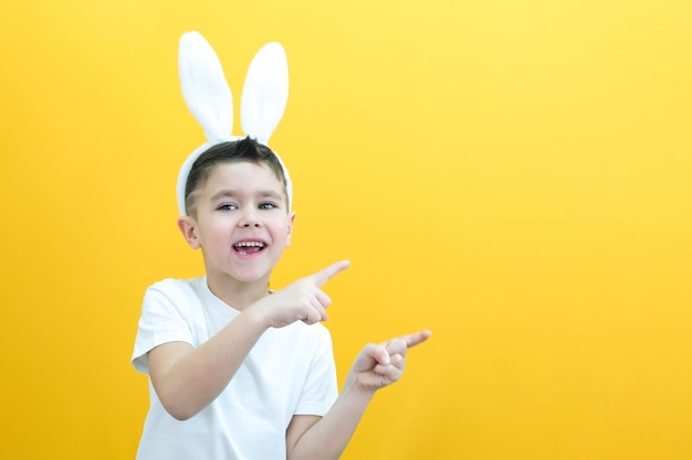 Ragazzo allegro con orecchie di coniglio sulla testa su sfondo giallo Bambino felice divertente punta il dito su uno spazio vuoto copia spazio per il layout del testo concetto di pasqua