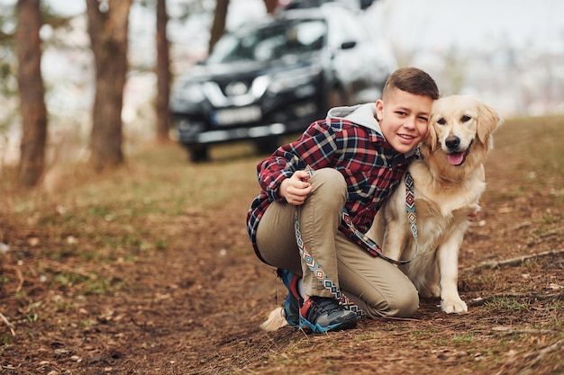 Ragazzo allegro in abiti casual seduto con il suo cane nella foresta contro la moderna macchina nera