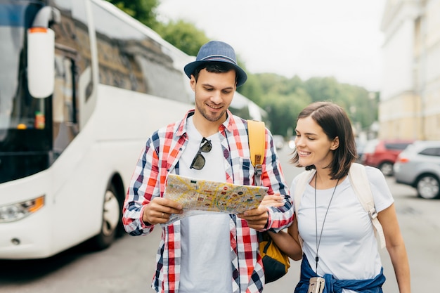 Ragazzo allegro in cappello e camicia, tenendo la mappa della città, guardando con un sorriso, in piedi vicino alla sua amica, camminando in città, esplorando nuovi spettacoli. Turisti maschi e femmine con guida della città.