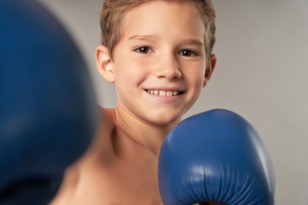 Ragazzo allegro in guantoni da boxe in piedi su sfondo grigio