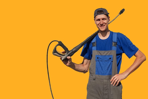 Ragazzo alto e magro con una pistola per un autolavaggio isolato su sfondo giallo Un lavoratore in tuta guarda la telecamera e sorride Sfondo o layout con spazio per la copia