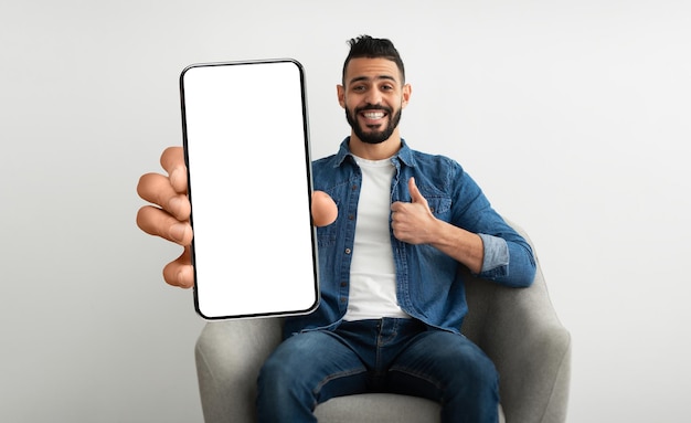 Ragazzo arabo allegro che si siede sulla sedia con il cellulare enorme con lo schermo vuoto bianco che mostra il pollice in su