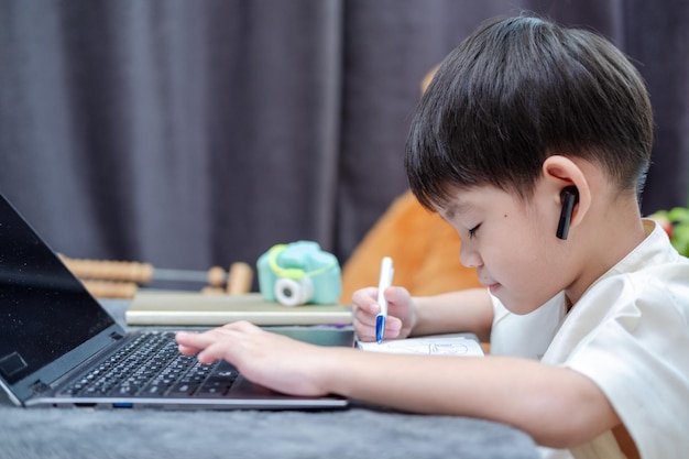 Ragazzo asiatico che ascolta l'audio e scrive i compiti mentre studia online sul laptop