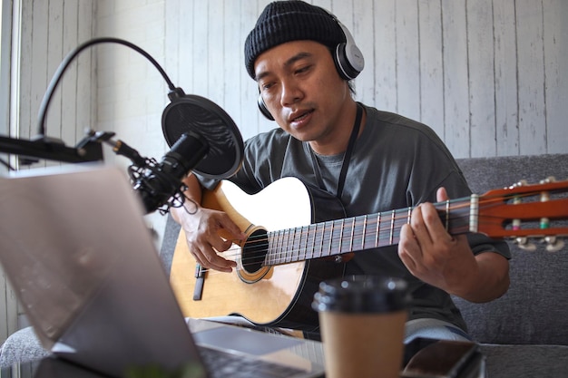 Ragazzo asiatico che suona il podcast audio di registrazione della chitarra in studio utilizzando microfono e laptop. Persone a