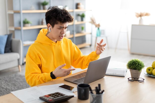 Ragazzo asiatico che usa gli auricolari da portare del computer portatile seduto alla scrivania