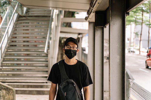 Ragazzo asiatico con gli occhiali e la maschera respiratoria nera si protegge per strada dal coronavirus