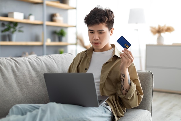 Ragazzo asiatico concentrato che mostra usando la carta di credito a casa