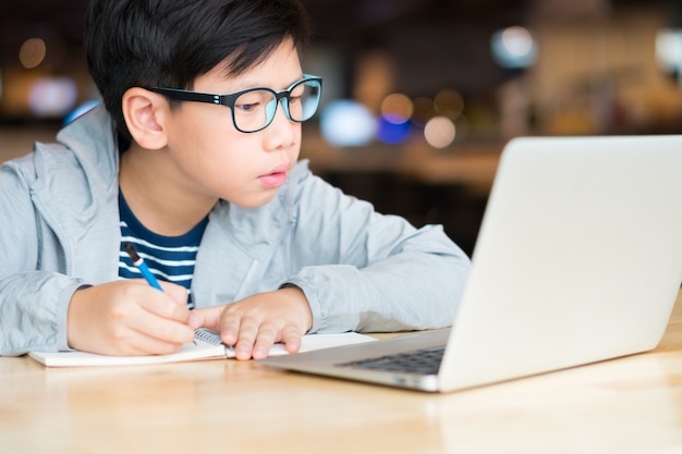 Ragazzo asiatico del preteen che sembra astuto che scrive e che per mezzo del computer portatile del computer che studia le lezioni online. Ricerca, studio e risoluzione di problemi con concentrazione. Apprendimento online e concetto di autoapprendimento.