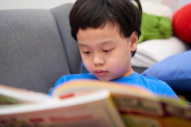 Ragazzo asiatico seduto sul divano con un libro da leggere e studiare da solo a casaÂ