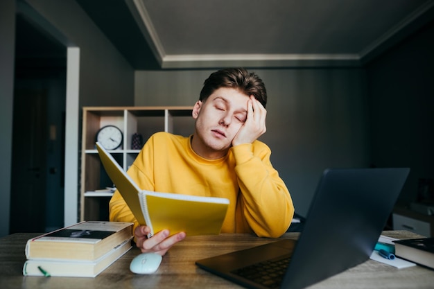 Ragazzo assonnato che studia a casa a un tavolo con libri e un laptop Quarantena