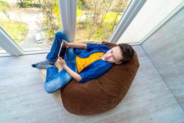 Ragazzo attraente che gioca nel tablet vicino alla finestra. Ragazzino che si siede sul pouf e gioca.