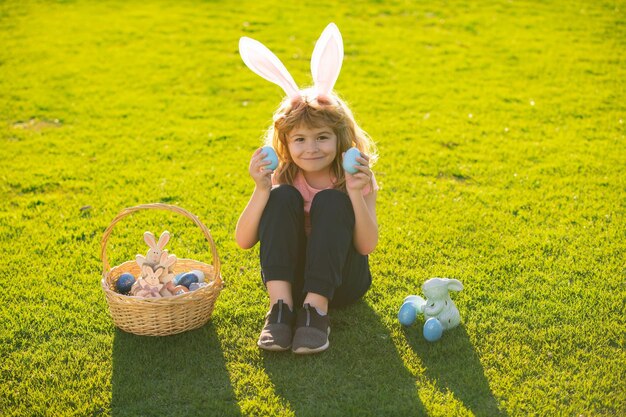 Ragazzo bambino in costume da coniglio con orecchie da coniglio che caccia uova di pasqua sull'erba nel parco primaverile ragazzo divertente ea