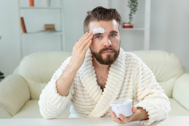 Ragazzo barbuto che applica la crema per il viso davanti allo specchio per la cura della pelle e la spa per il concetto dell'uomo