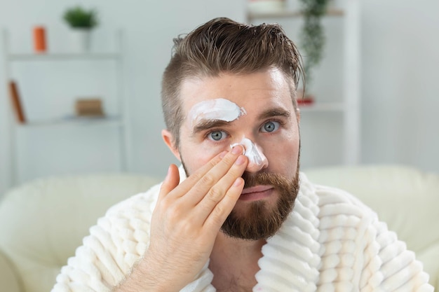 Ragazzo barbuto che si applica una crema antirughe sul viso. Cura della pelle e spa per il concetto di uomo.