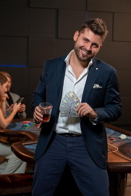 Ragazzo barbuto che vince nel poker in piedi con un bicchiere di bevanda e banconote nel casinò