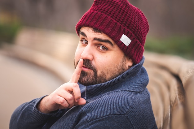 Ragazzo barbuto in un cappello di lana e un maglione seduto su una panchina nel parco Guardando la telecamera e chiedendo silenzio