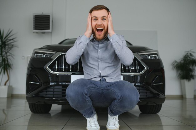 Ragazzo bello emotivo che alza la mano e sorride alla macchina fotografica Uomo ricco che compra auto al salone della concessionaria di lusso