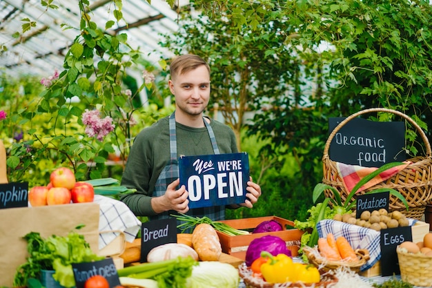 Ragazzo bello in grembiule con cartello aperto nel mercato degli alimenti biologici che accoglie le persone