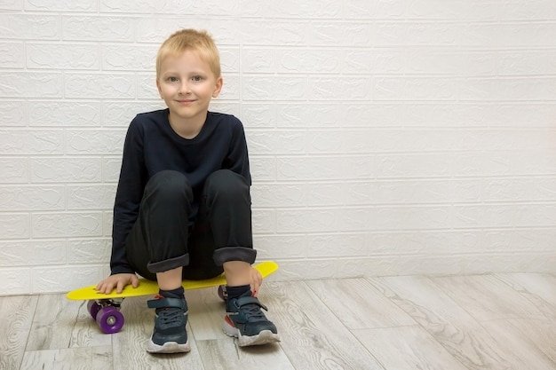 Ragazzo biondo carino sorridente stanco seduto su uno skateboard dopo l'allenamento contro un muro bianco