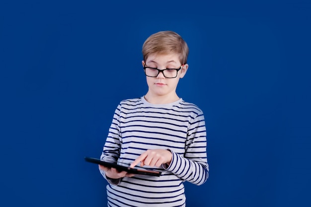 Ragazzo biondo del bambino con i grandi vetri che giocano con la compressa sulla parete blu. Concetto di educazione.