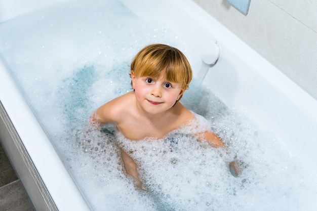 Ragazzo biondo dolce del bambino che cattura il bagno nella vasca da bagno e che gioca nella schiuma di sapone.