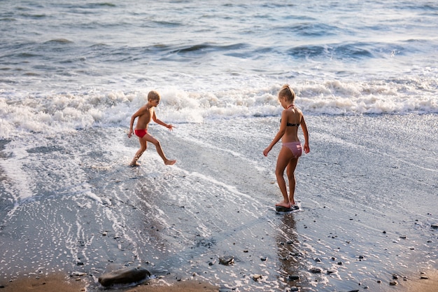 Ragazzo biondo in costume da bagno rosso e ragazza che giocano e si divertono sulla spiaggia sulla riva del mare blu nelle vacanze estive al tempo di giorno.
