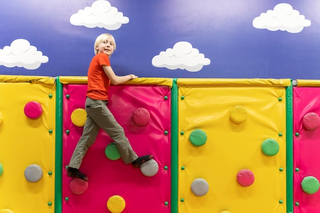 Ragazzo biondo nella sala giochi per bambini sulla parete da arrampicata Il bambino si diverte nel centro di intrattenimento