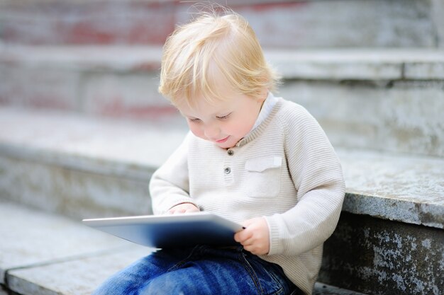 Ragazzo biondo sveglio del bambino che gioca con una compressa digitale all&#39;aperto