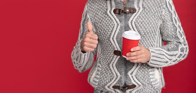 Ragazzo brizzolato in maglione e cappello con paraorecchie beve caffè su sfondo rosso che mostra il pollice al mattino
