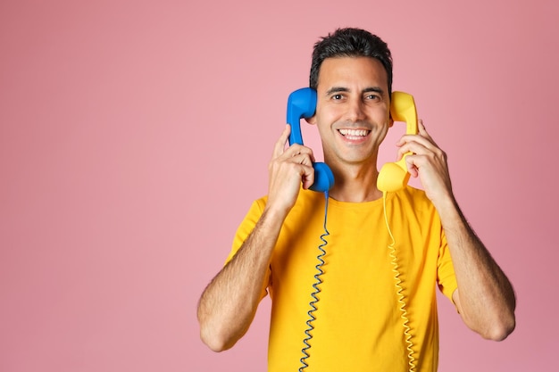 Ragazzo bruna sorridente in maglietta gialla che tiene due telefoni blu e gialli vintage nelle orecchie su sfondo rosa