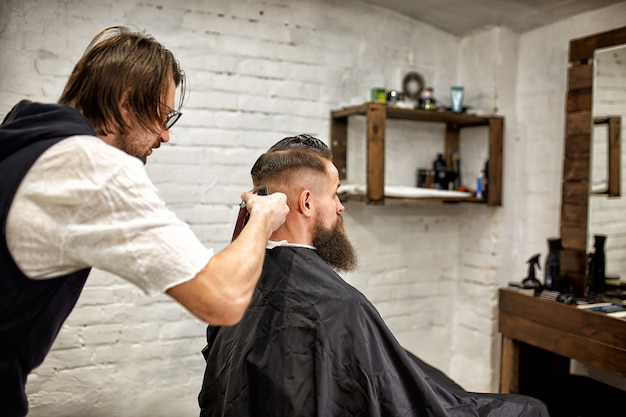 Ragazzo brutale nel moderno Barber Shop. Il parrucchiere fa acconciatura un uomo con la barba lunga. Il maestro parrucchiere fa acconciatura con una tosatrice