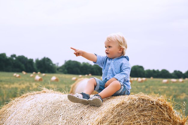 Ragazzo carino bambino seduto su pagliai al campo di grano Bambino felice al giorno d'estate sulla natura all'aperto Sfondo dell'infanzia della famiglia