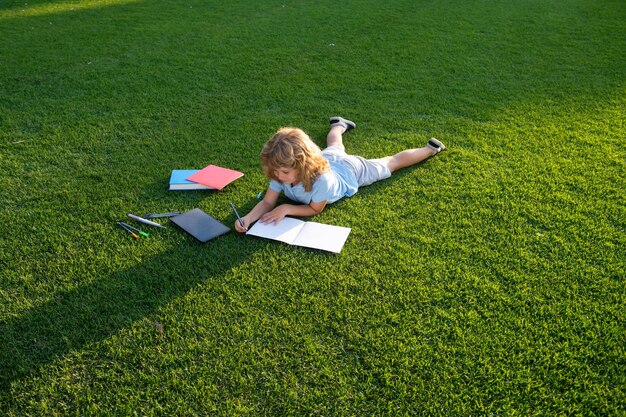 Ragazzo carino che scrive sul taccuino posa sull'erba Bambino che legge un libro nel parco estivo Concetto di bambini