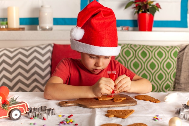 Ragazzo carino con cappello di Babbo Natale decora con cura il pan di zenzero