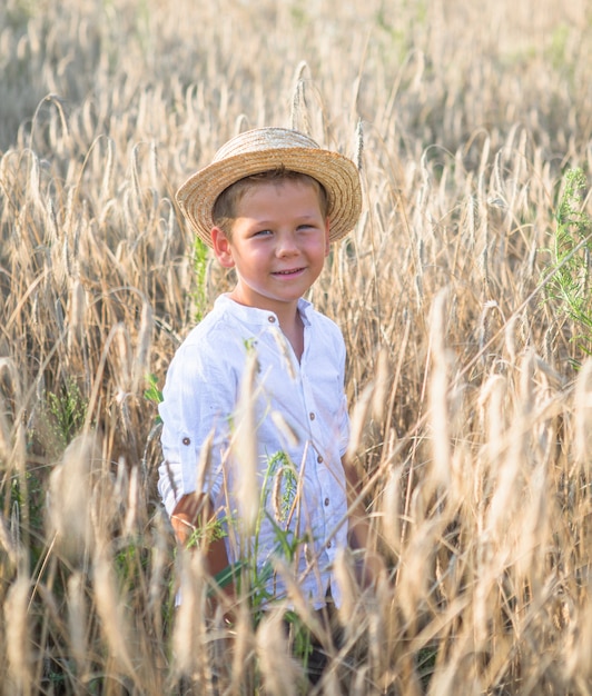 Ragazzo carino con cappello di paglia con margherite in mano che camminano sul campo di grano di segale