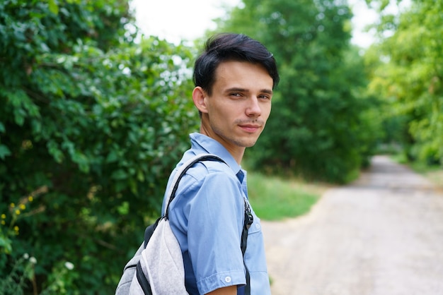 Ragazzo carino con uno zaino in piedi sulla strada in campagna, giovane caucasico in camicia blu viaggia h...