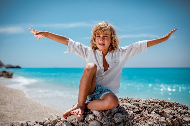 Ragazzo carino è seduto sulla roccia del mare sulla spiaggia e sorride guardando la fotocamera. Le sue braccia sono spalancate.