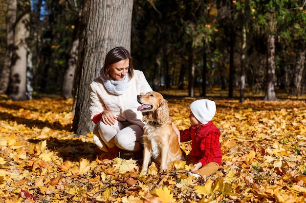 Ragazzo carino, felice, bianco in camicia rossa che sorride e gioca con il cane tra le foglie gialle. Piccolo bambino che si diverte con sua madre nel parco autunnale. Concetto di amicizia tra bambini e animali domestici, famiglia felice