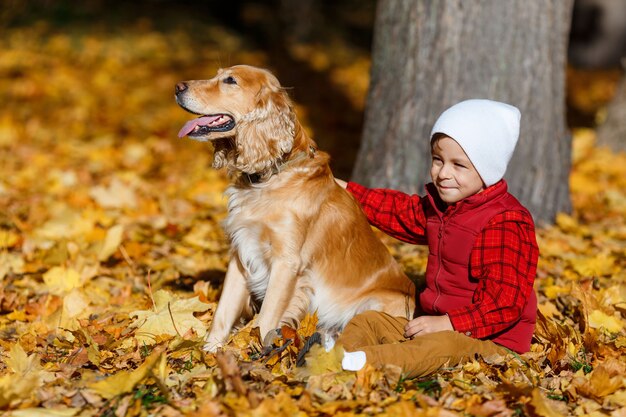 Ragazzo carino, felice, bianco in camicia rossa che sorride e gioca con il cane tra le foglie gialle. Piccolo bambino che si diverte nel parco autunnale. Concetto di amicizia tra bambini e animali domestici, famiglia felice