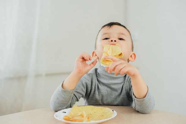 Ragazzo carino mangiare deliziose frittelle seduto a un tavolo su uno sfondo chiaro carino