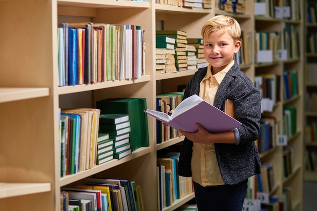 ragazzo carino scuola sorridente durante la lettura di un libro in biblioteca, stare in piedi guardando la fotocamera