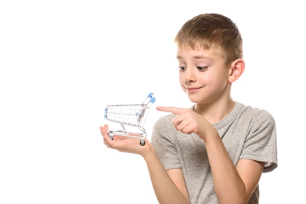 Ragazzo carino tiene in mano il piccolo carrello della spesa in metallo e lo indica con il dito indice. Isolare su sfondo bianco. Concetto di shopping.