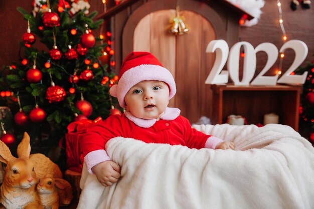 Ragazzo carino vestito da Babbo Natale si siede vicino all'albero di Natale. merci per bambini e vacanze. Concetto di Capodanno invernale. natale 2022