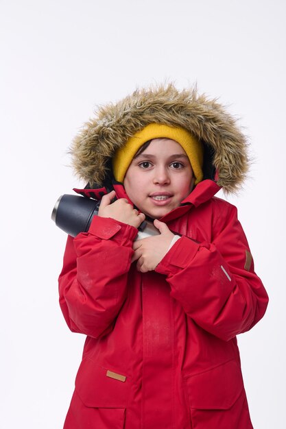 Ragazzo carino viaggiatore avventuriero in età scolare che si scalda abbracciando il thermos, tremando dal freddo guardando la telecamera isolata su sfondo bianco con spazio di copia per gli annunci. Tempo libero invernale, concetto di vacanze